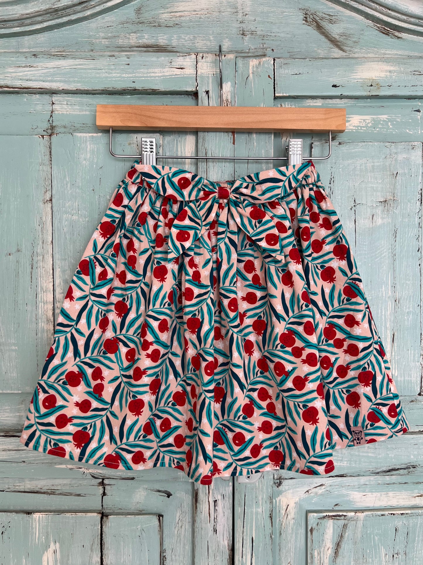 soft organic cotton skirt for girls age 2 to 8 years old. flowery print with a soft pink base and green and red pomegranate, featuring a wavy style,flowy design, elastic waistband and matching fabric decorative bow.