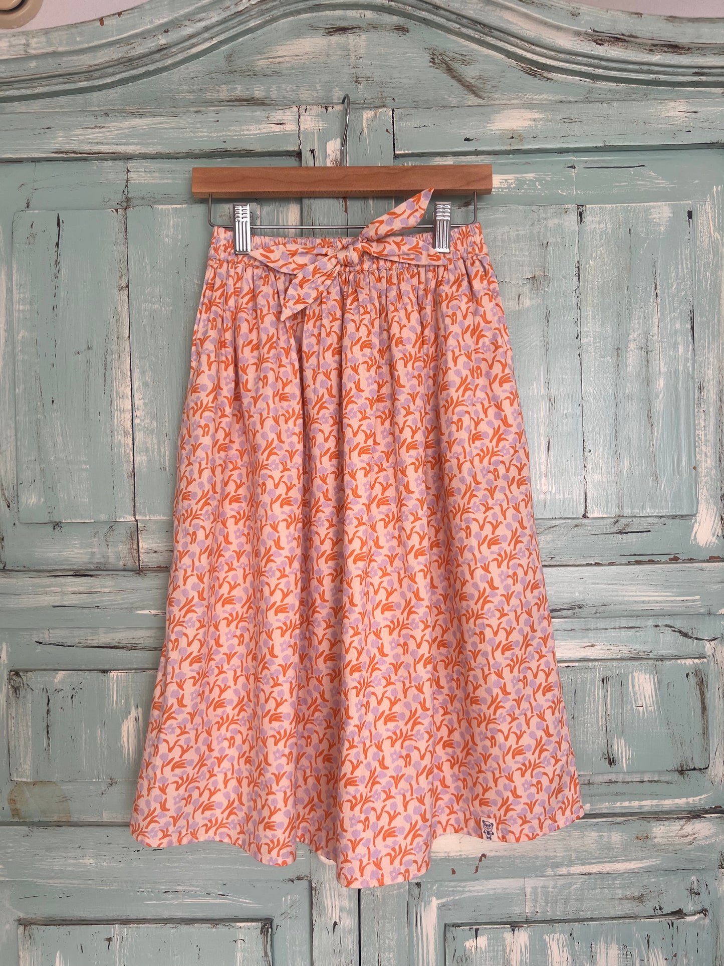 organic cotton skirt for girls aged 6 to 8 years old, featuring a soft pink base with an orange and violet botanical print, a gathered waist, and a matching fabric tie.